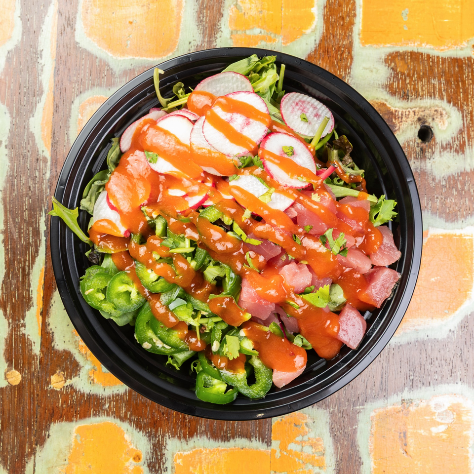 A vibrant poke bowl with tuna, radishes, jalapeños, and a spicy orange sauce on a rustic wooden table.