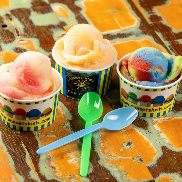 Three cups of Sunset Slush Italian ice in various flavors and colors on a rustic wooden table.